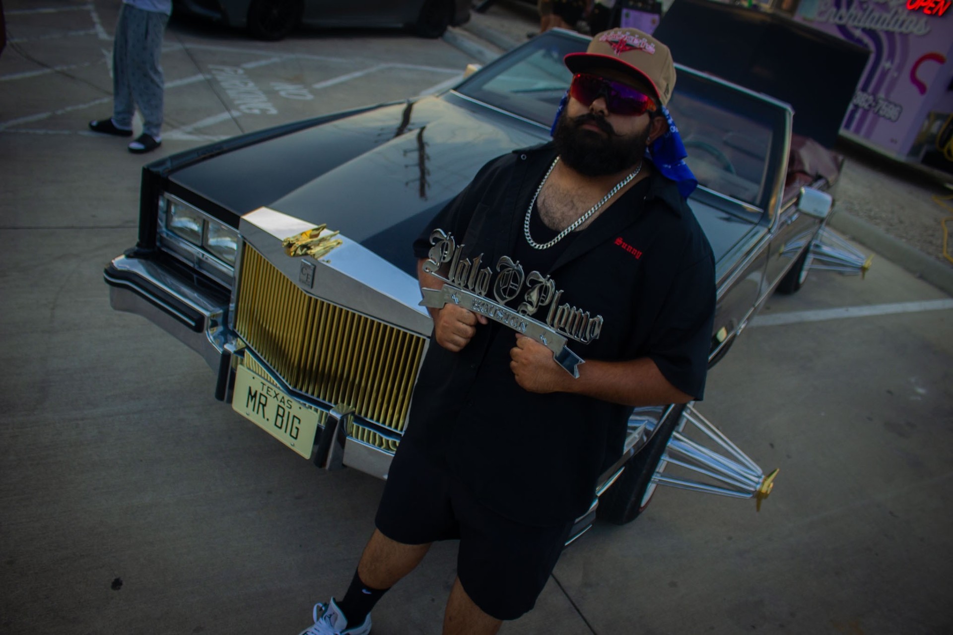 Chrome Plata O Plomo Houston club plaque held in front of a gold-grille Cadillac
