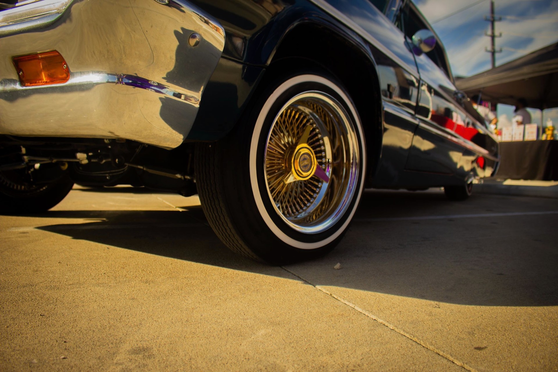 Gold and chrome wire wheel with whitewall tire under a black '64 Impala